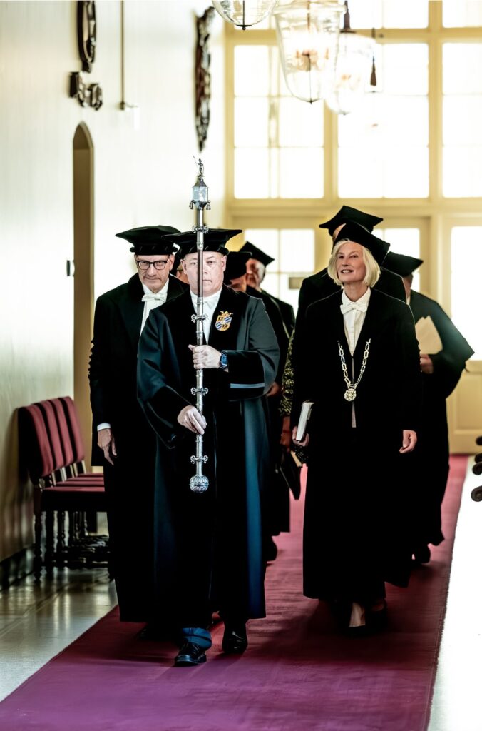 Academische stoet met hoogleraren in traditionele toga en baret loopt door de gangen van de Rijksuniversiteit Groningen tijdens een promotieplechtigheid, met de pedel voorop die de universitaire scepter draagt.